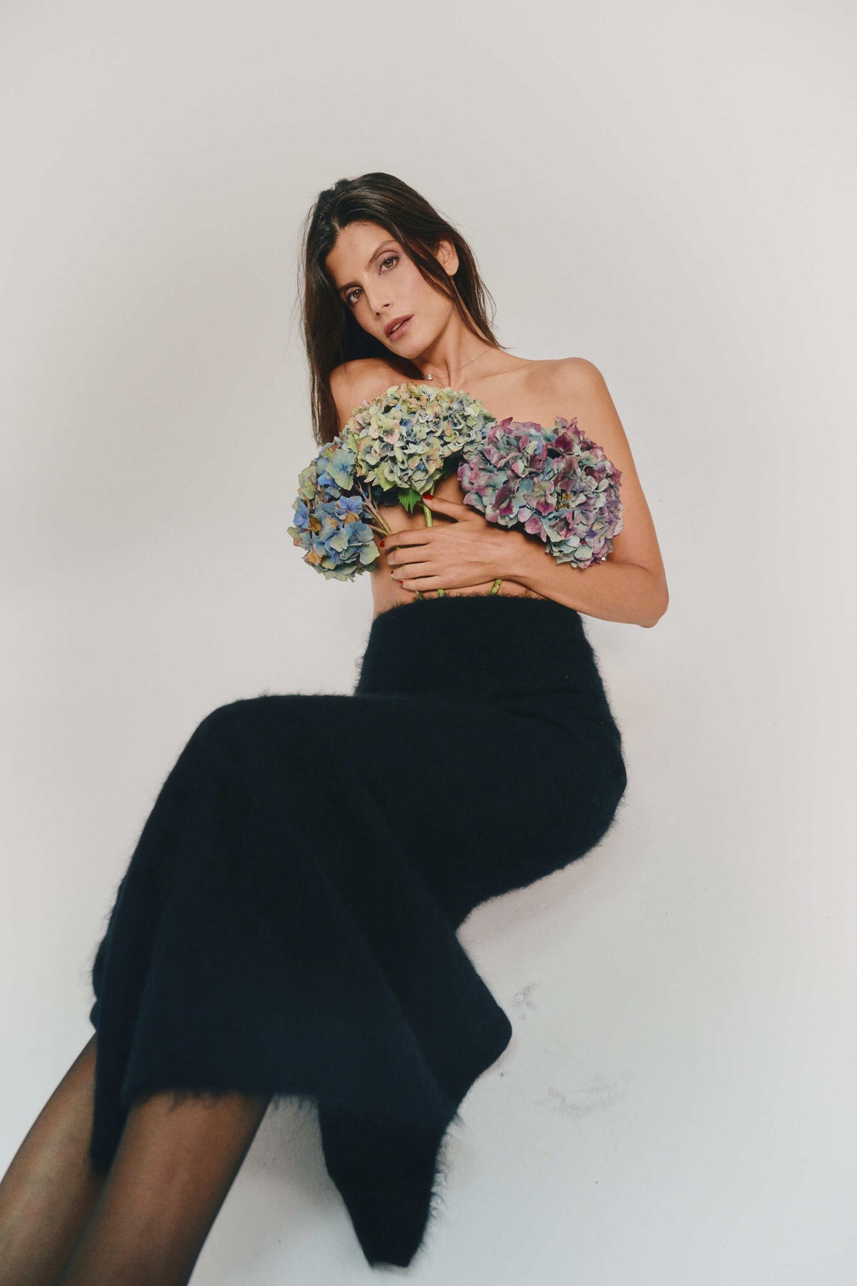 A woman poses in a long black cashmere skirt while holding a large bouquet of blue and purple hydrangeas against a plain white studio background.