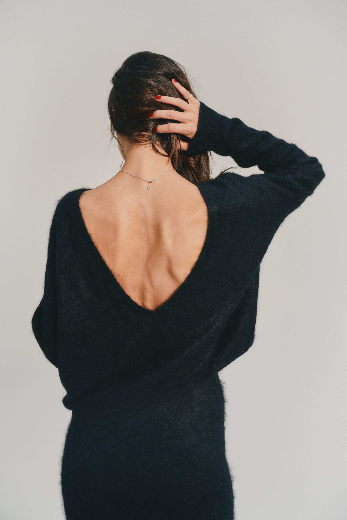 A woman shown from behind wearing a black brushed cashmere knitwear with a very large open back, posed against a plain light gray background.