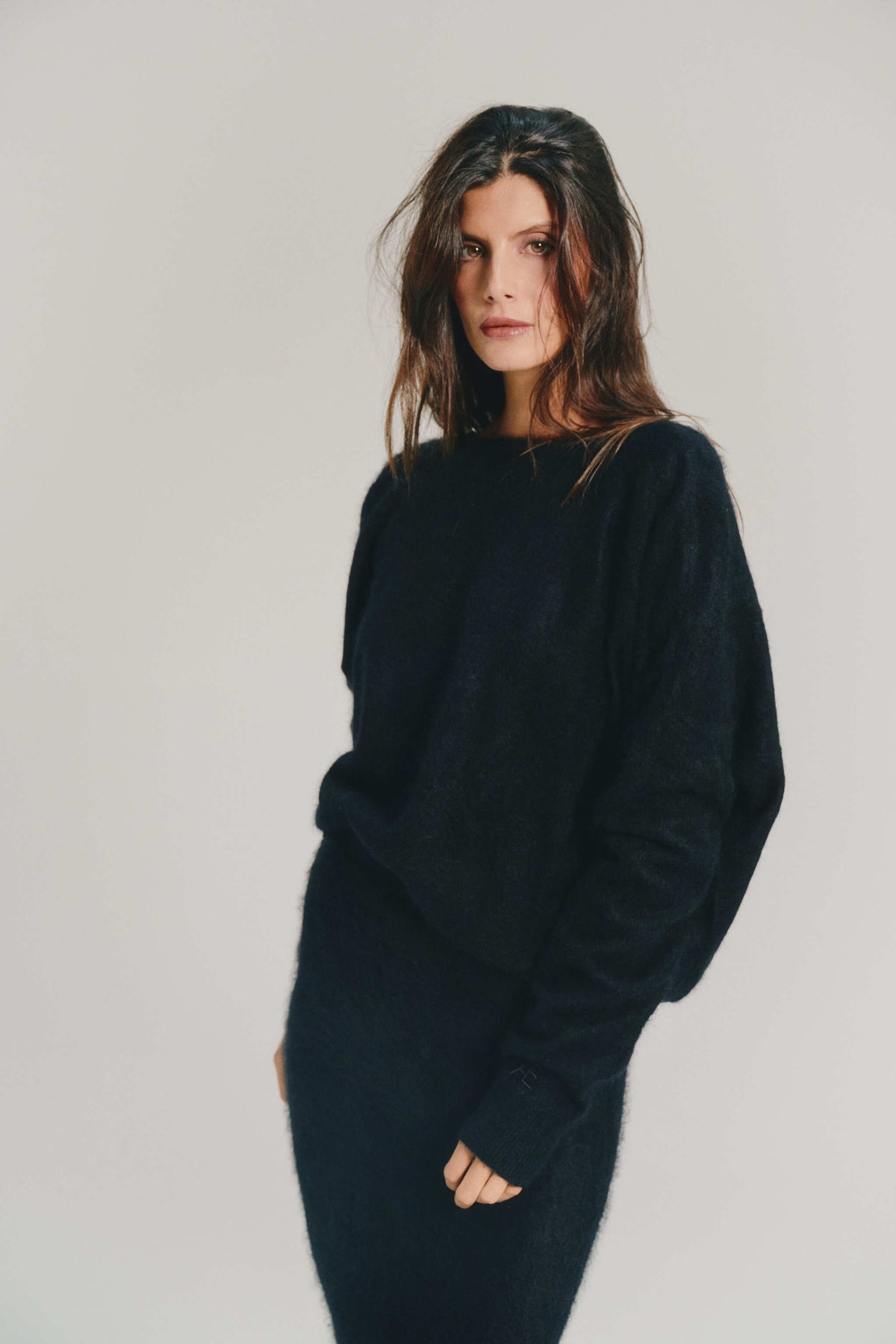A woman with dark hair wearing a black knitwear wearing a cashmere knitwear sweater and matching black skirt, posing against a light gray studio background.