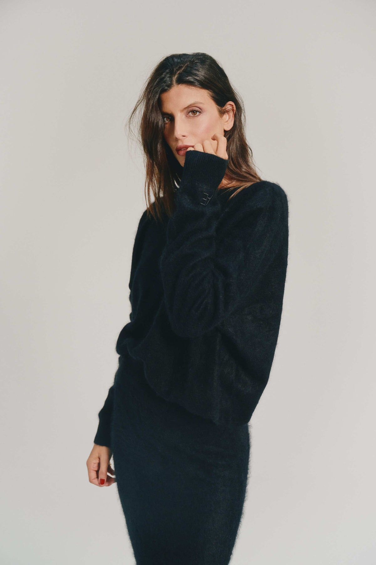 A woman modeling a black cashmere knitwear and matching skirt against a plain light gray studio background.