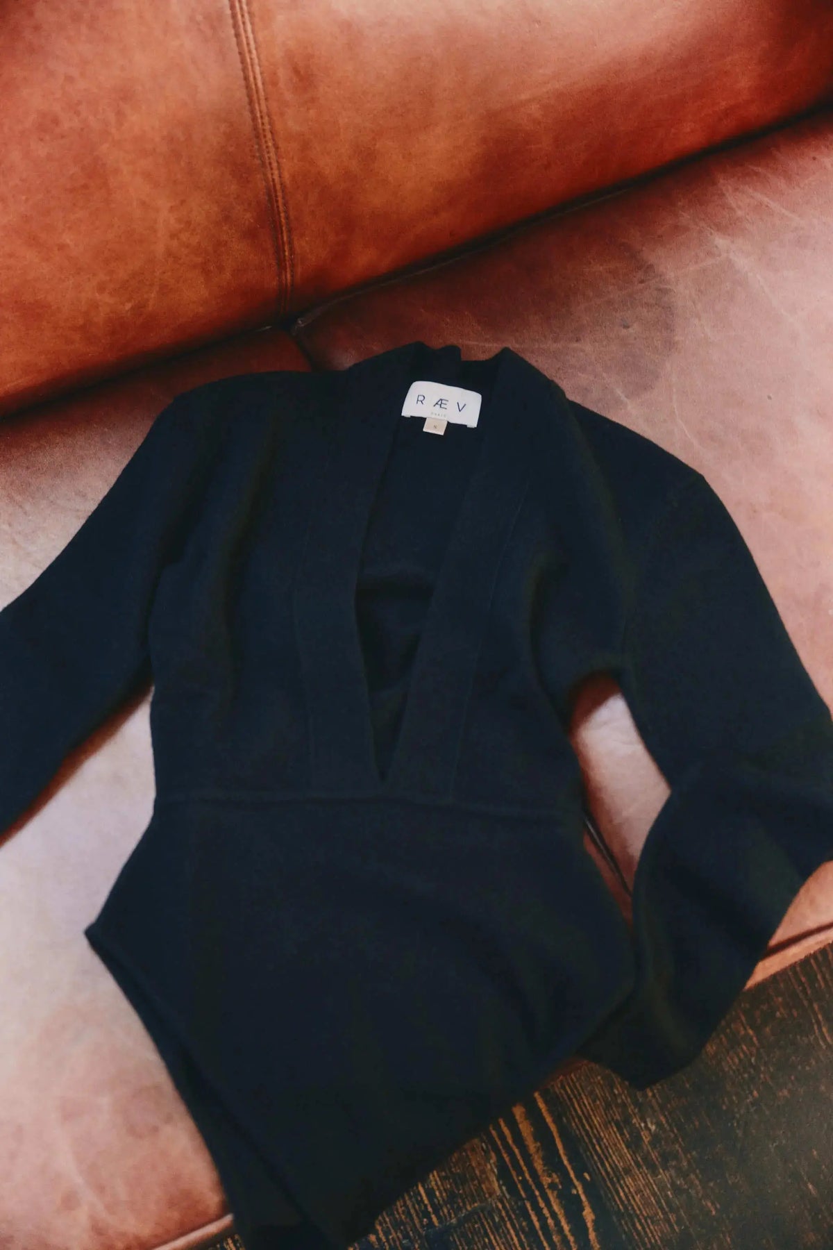 A black long-sleeve cashmere bodysuit  with a deep V-neckline, laid flat on a brown leather sofa. RAEV label is visible at the collar.
