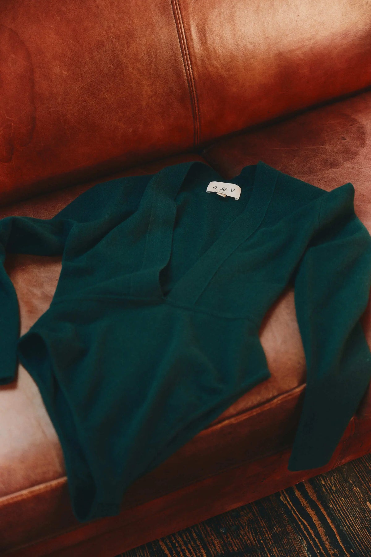 A dark green long-sleeve cashmere bodysuit laid flat on a brown leather couch, with RAEV label visible.