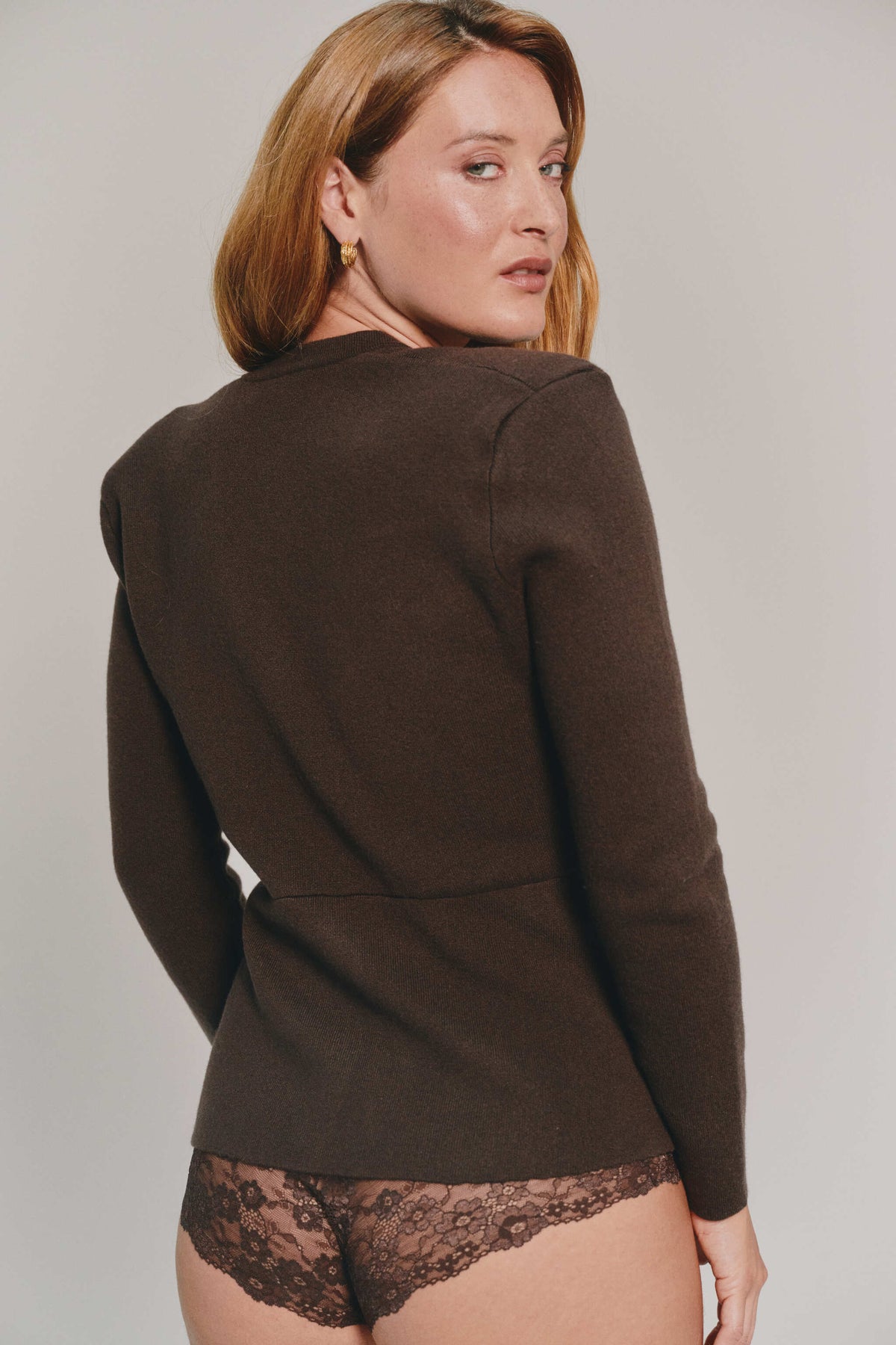 A woman shown from the back wearing a dark brown cashmere cardigan and brown lace underwear, photographed against a plain light gray studio background.