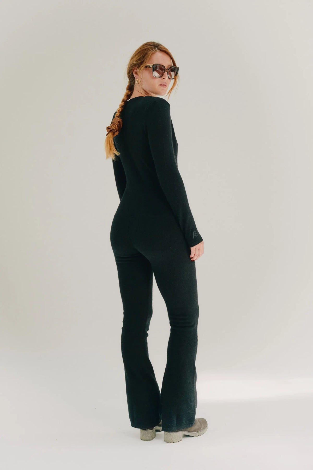 A woman with a long blonde braid and sunglasses, wearing a black long-sleeve flared jumpsuit in cashmere, standing in profile against a light gray studio background.
