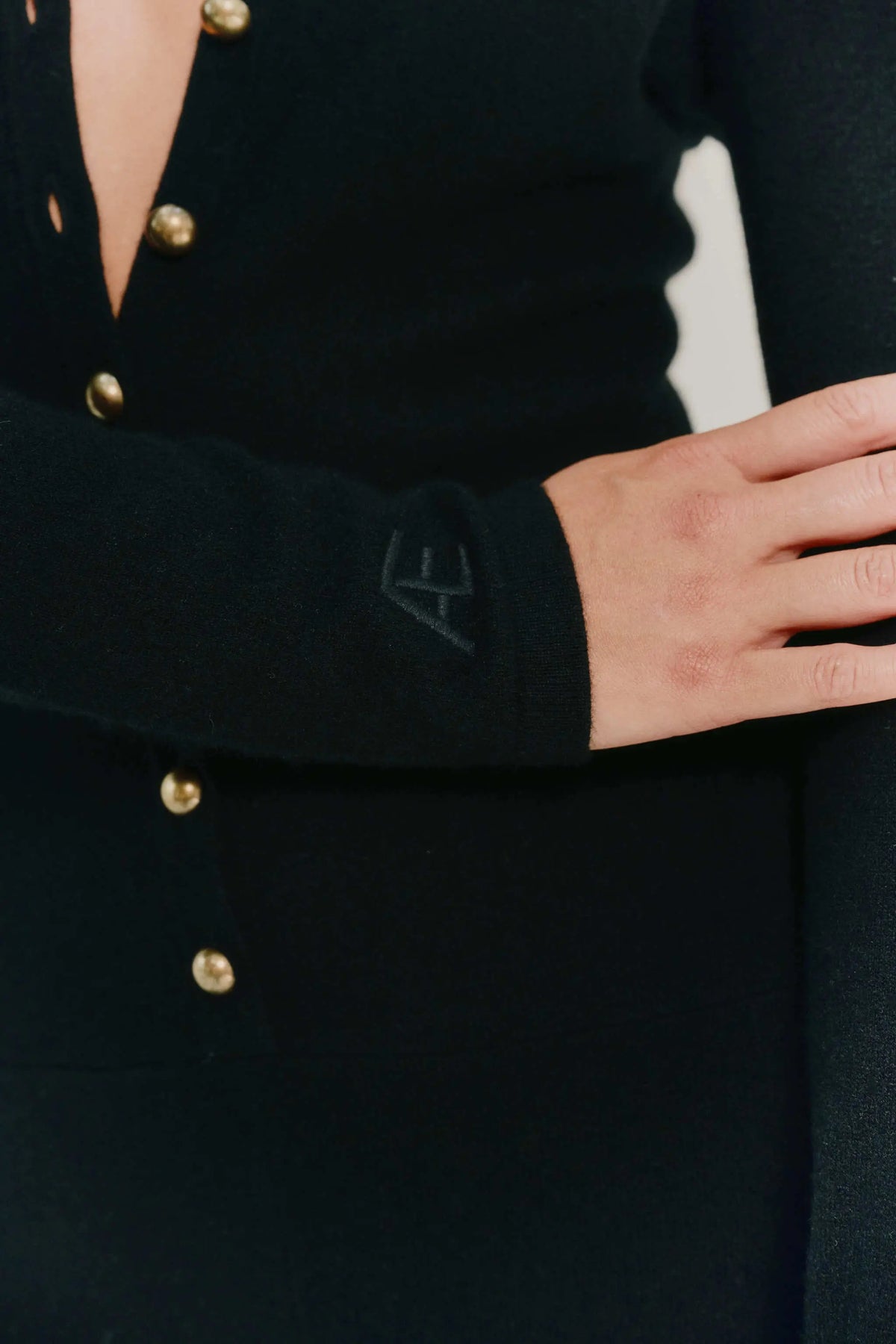 Close-up of a person wearing a black long-sleeve cashmere jumpsuit with gold buttons and a tonal embroidered monogram on the cuff.