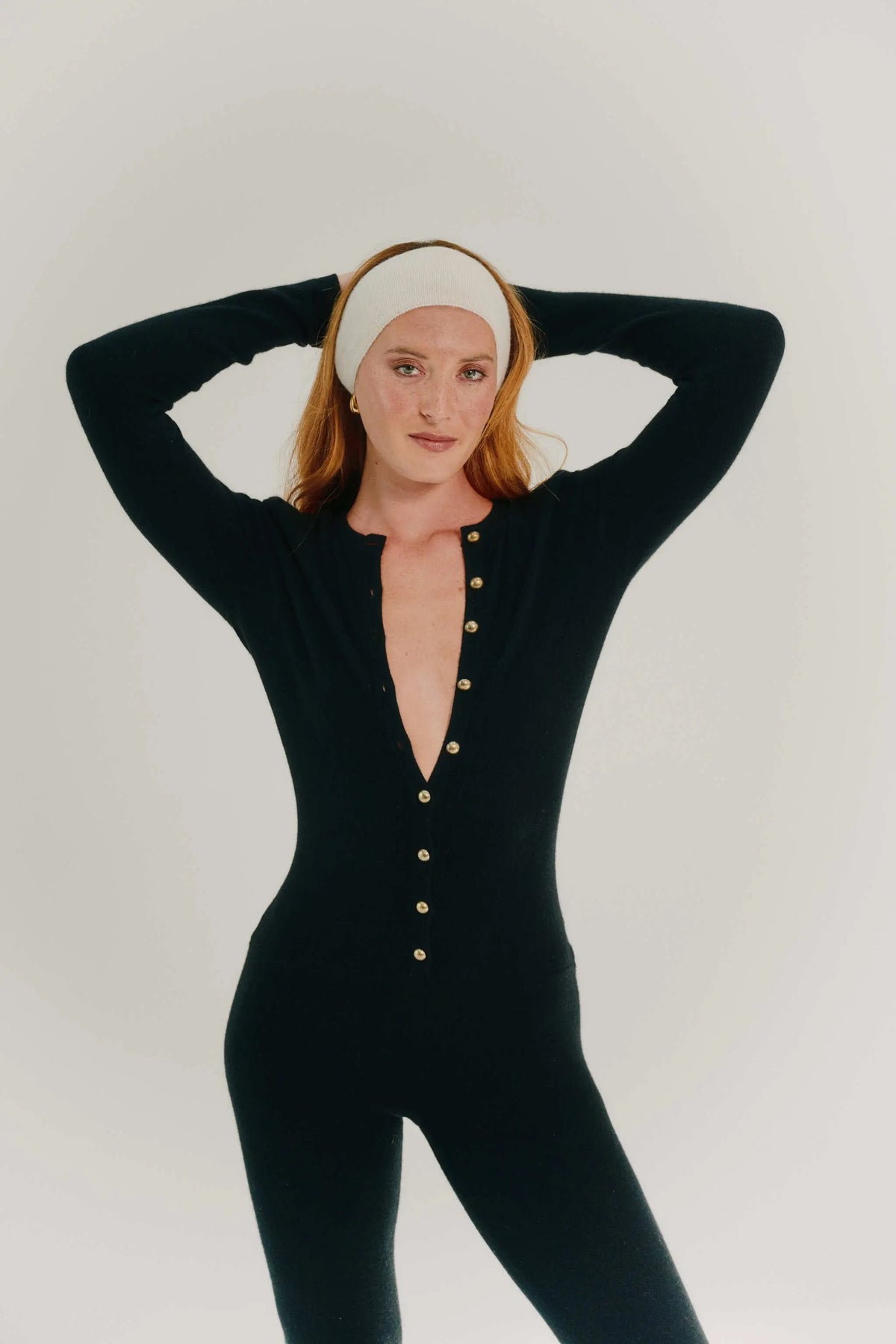 A woman modeling a black long-sleeve cashmere bodysuit with gold buttons down the front, styled with a white headband, against a light gray studio background.