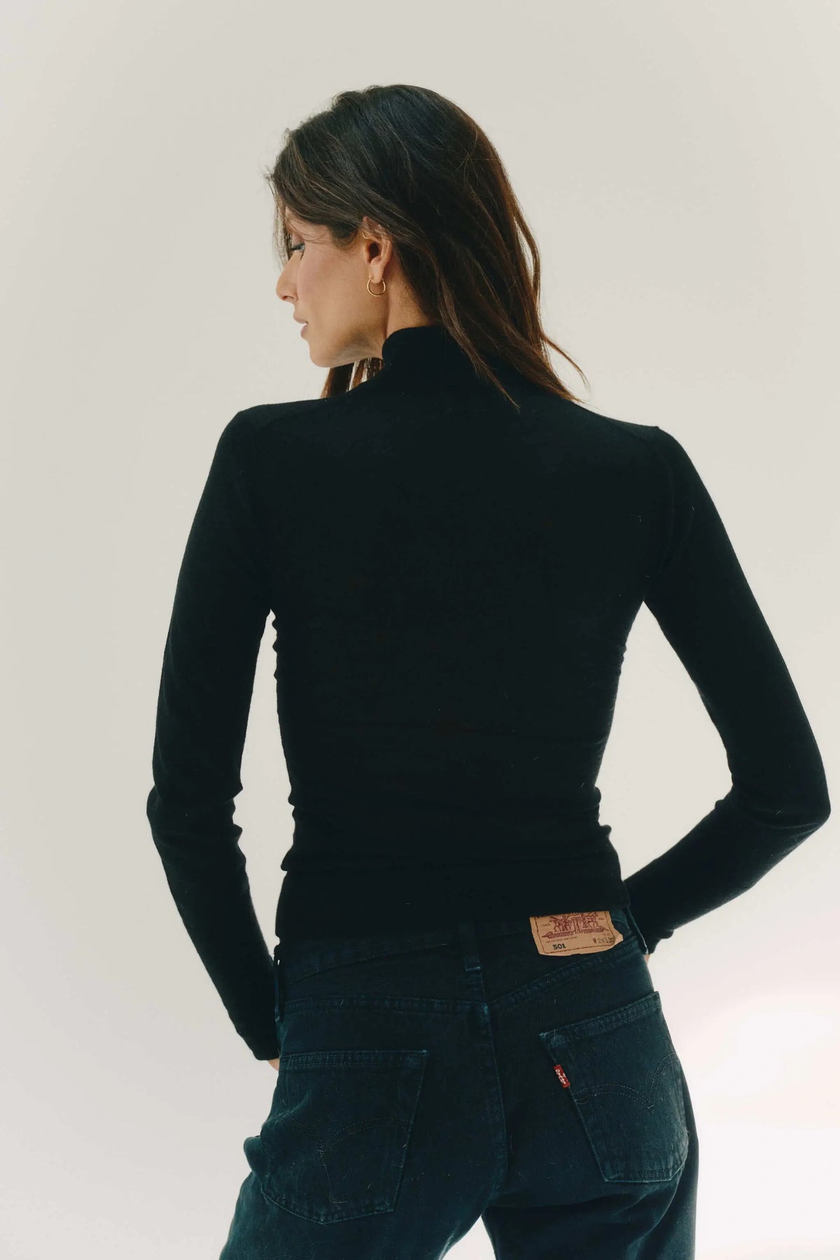 A woman shown from the back wearing a fitted black long-sleeve mock-neck cashmere top and dark blue jeans against a plain light background.