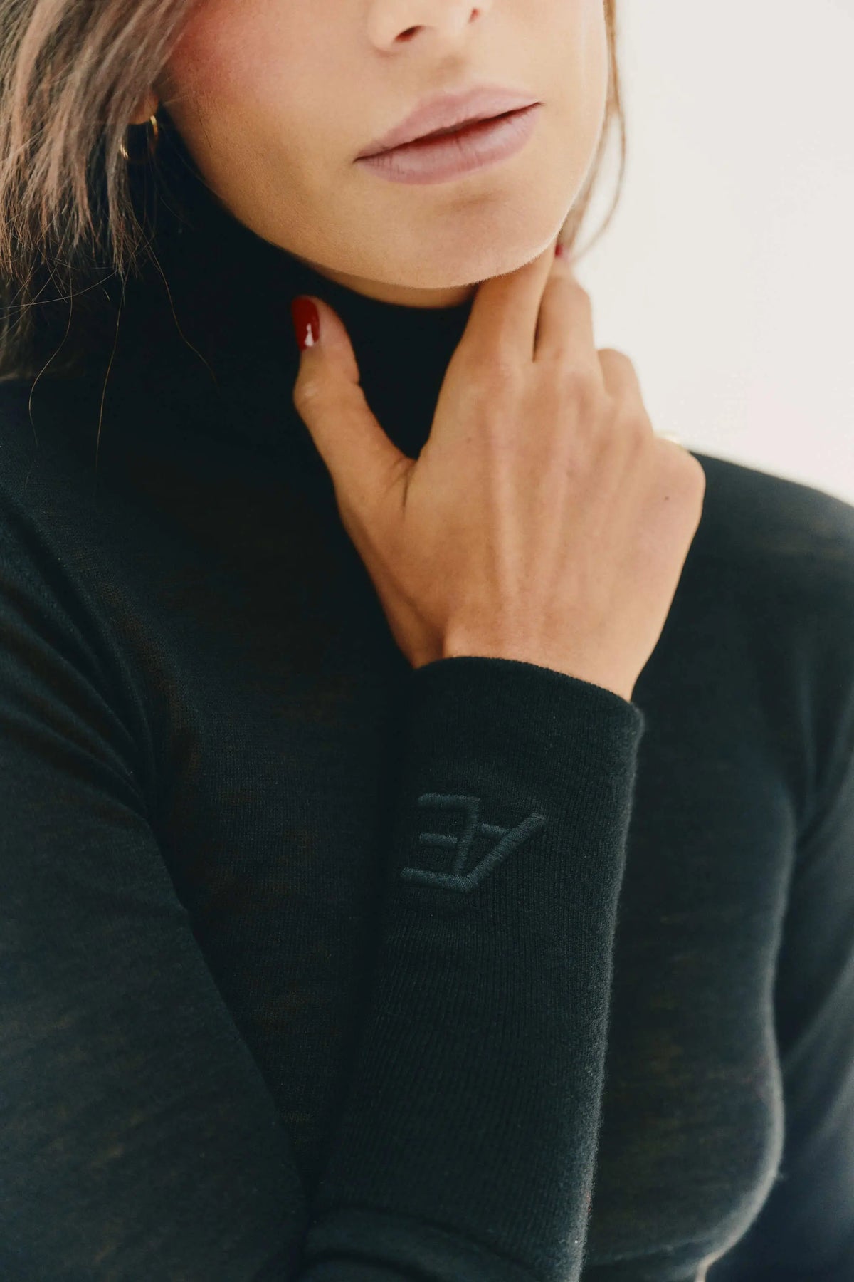 Close-up of a woman wearing a black ribbed turtleneck cashmere sweater with a tonal embroidered logo on the cuff, set against a bright off-white background.