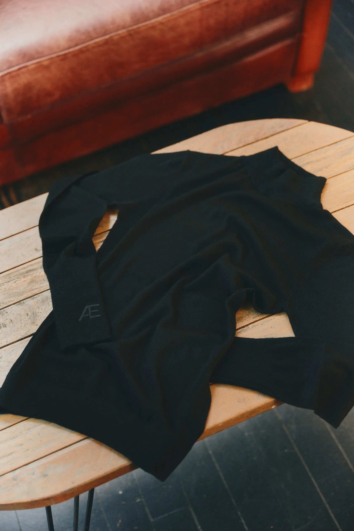 A black long-sleeve knit cashmere top with a small gray monogram logo on the cuff, draped on a light wood slatted table with a brown leather sofa in the background.