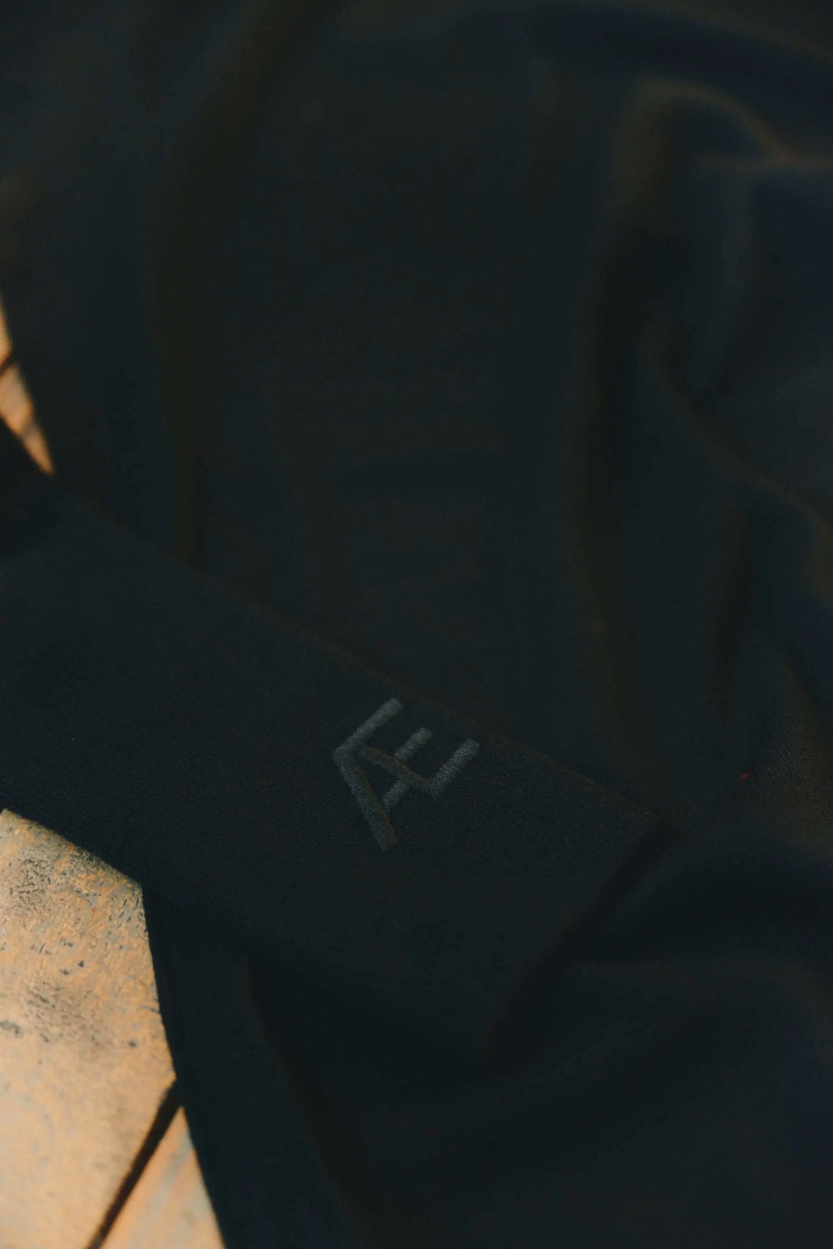 A close-up shot of a black cashmere top with a tonal black embroidered logo, resting on a textured wood surface.