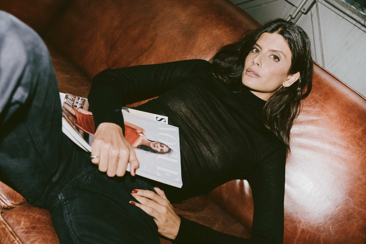 A woman with dark hair lies on a brown leather couch, wearing a transparent black long-sleeve turtleneck in cashmere and black jeans, holding a magazine.