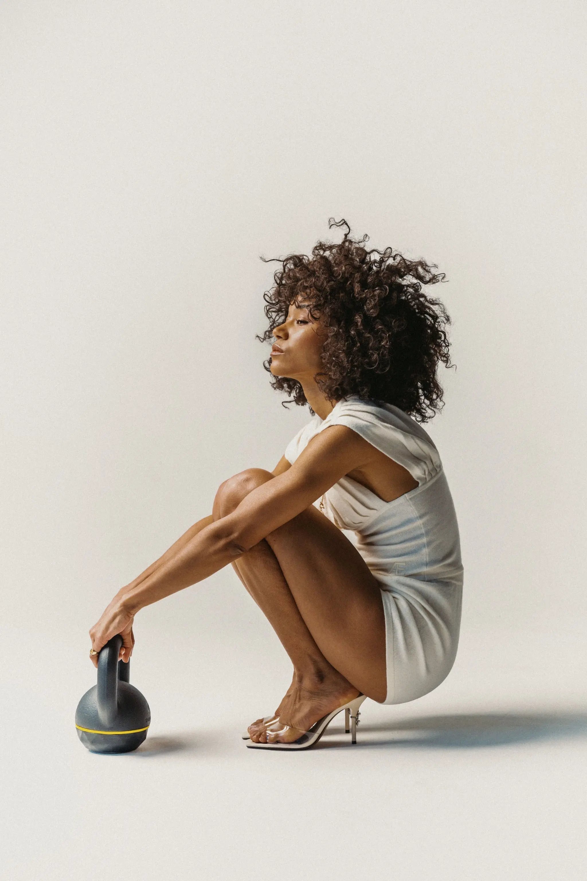Woman in a white outfit sitting with a kettlebell on a plain background