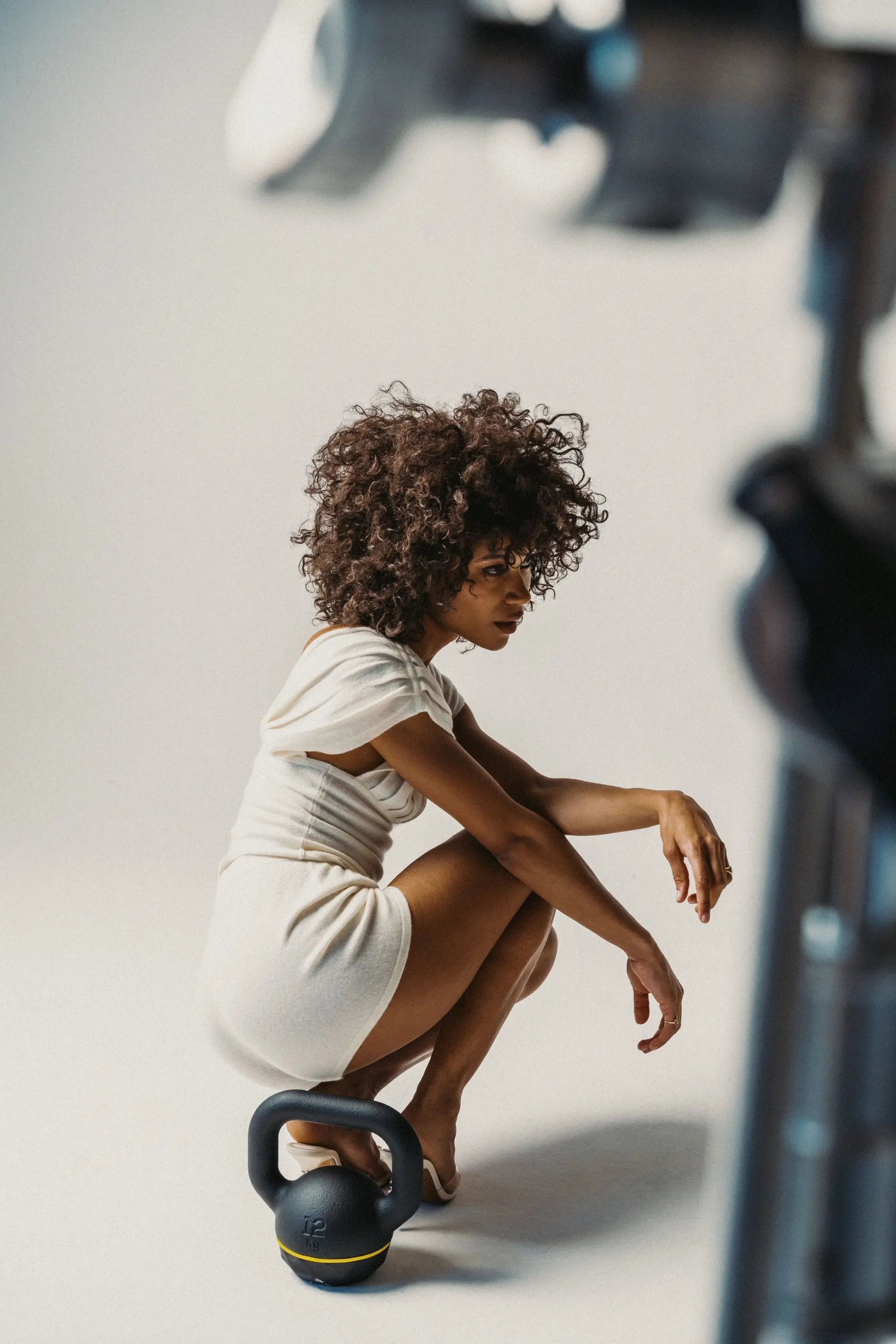 Woman in white outfit sitting on a kettlebell with a camera setup in the foreground