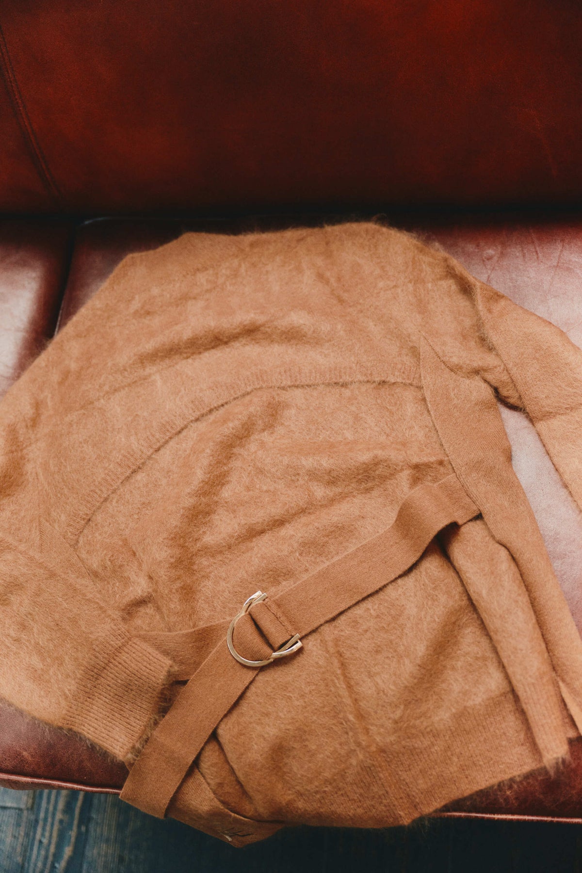 A camel cashmere backless sweater with a matching belt and silver-tone ring buckle, laid flat on a brown leather chair.