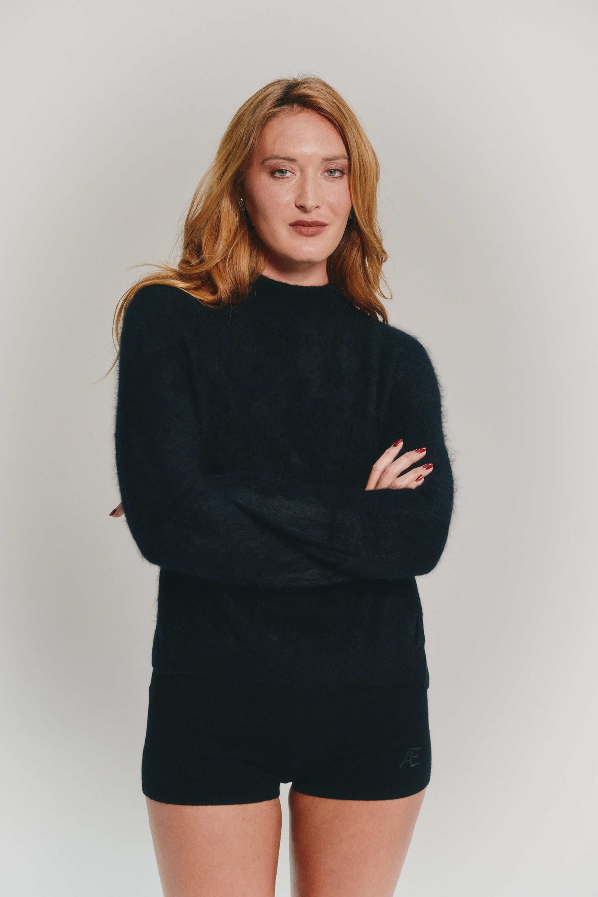 A woman with long wavy light red hair wearing a black cashmere mock-neck sweater and matching black cashmere shorts, standing with arms crossed against a light gray studio background.