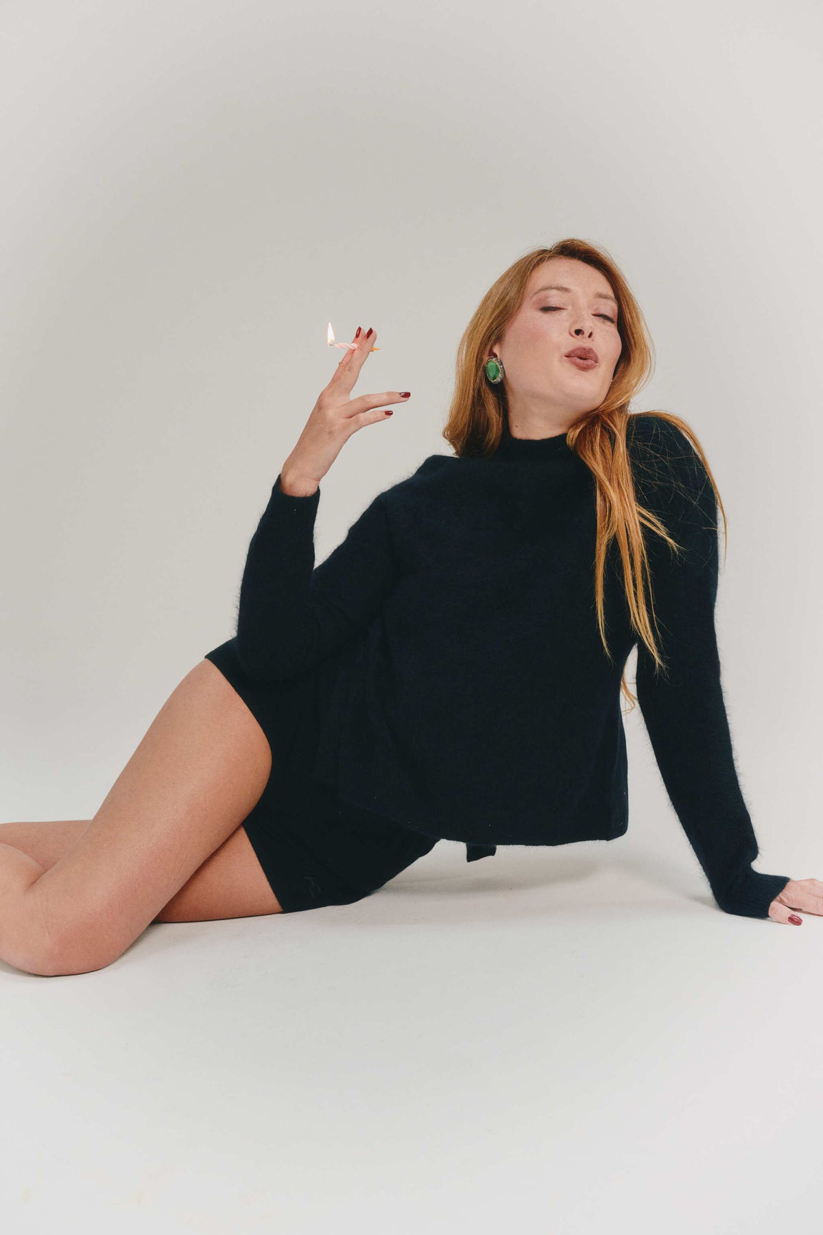 A woman with long red hair poses on a white studio floor wearing a black long-sleeve cashmere knirwear. She is holding a small white object near her face, with one hand resting on the floor.