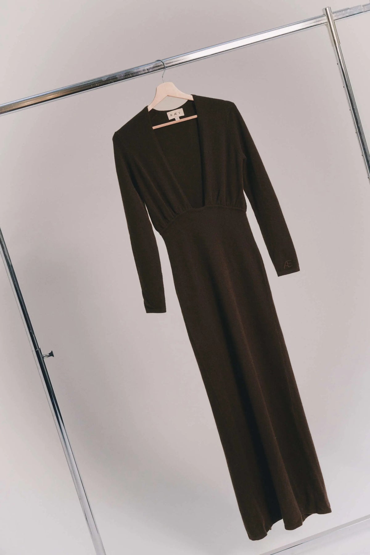 A dark brown long-sleeve knit maxi cashmere dress hanging on a wooden hanger from a metal clothing rack, shown against a light gray wall.