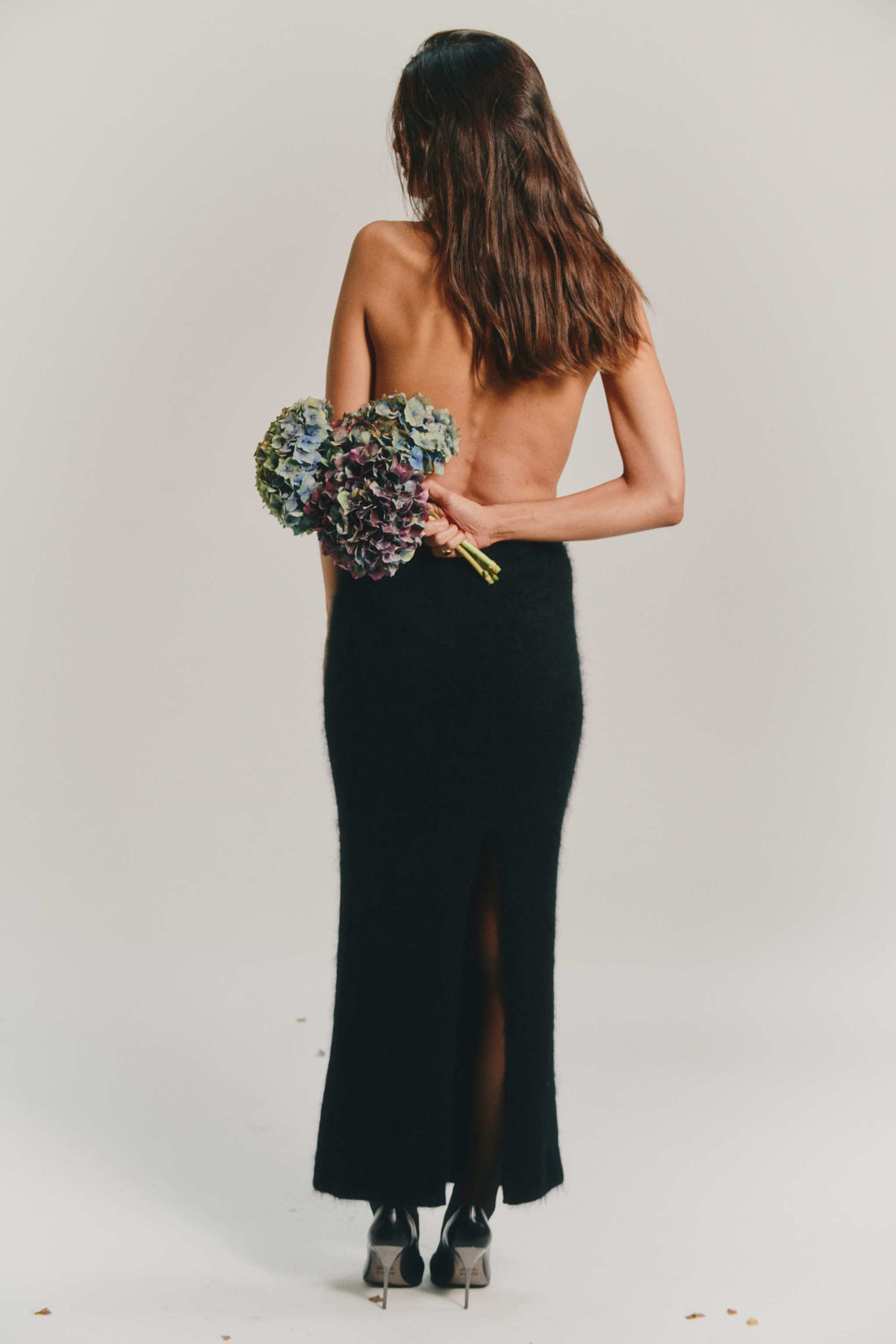 A woman photographed from behind wearing a long black cashmere skirt with a high slit, holding a bouquet of blue and purple flowers against a plain light gray studio background.