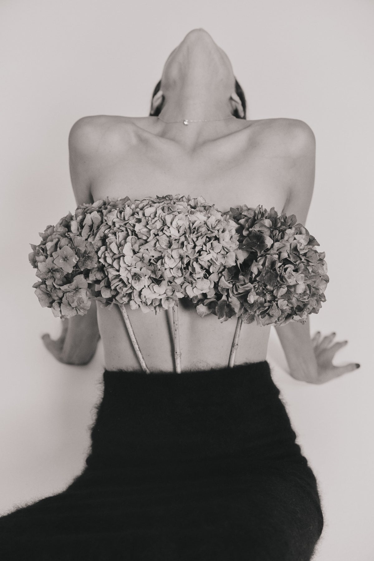 A black and white artistic photograph of a model from the front, wearing a black brushed cashmere skirt with a massive, structured bouquet of hydrangeas across the bust.