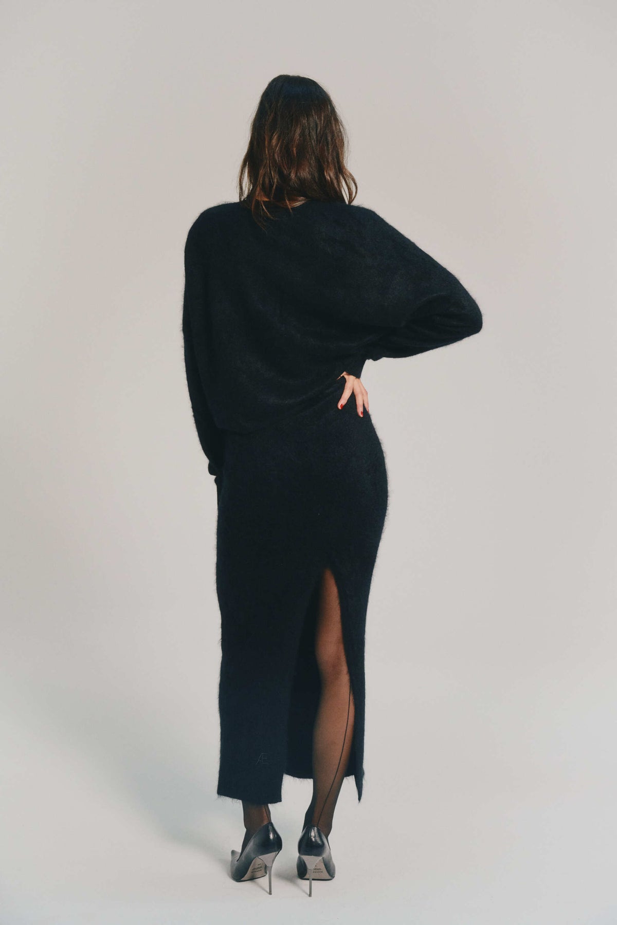 A woman is shown from behind wearing a long black cashmere skirt with a thigh-high slit, paired with black tights and black high-heeled shoes, against a light gray studio background.