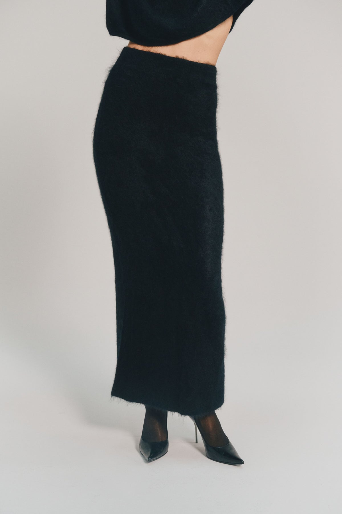A model is shown from the waist down wearing a long black cashmere skirt paired with black pointed-toe heels against a plain light gray studio background.