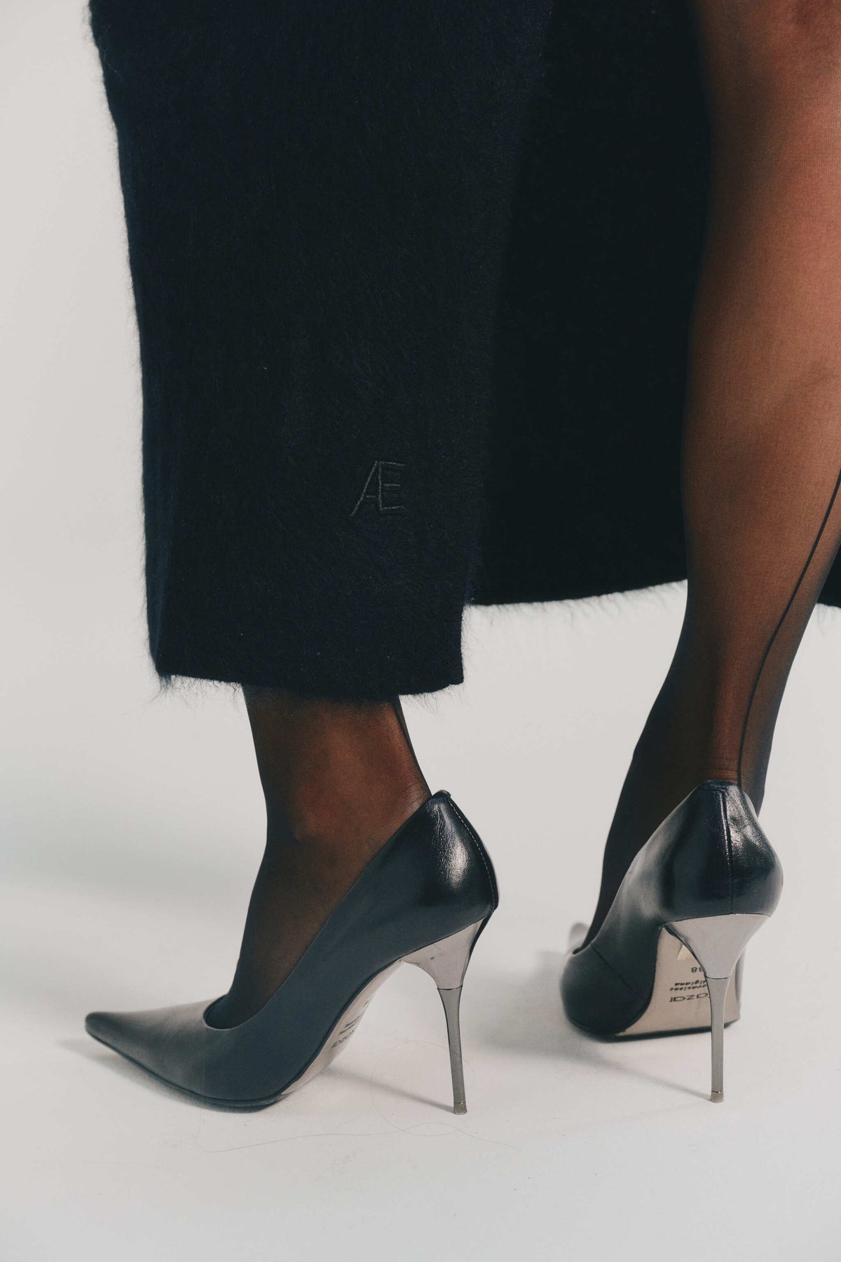 A close-up of a person wearing black pointed-toe stiletto pumps with silver heels, paired with sheer black tights and dark cashmere skirt, photographed against a clean white studio background.