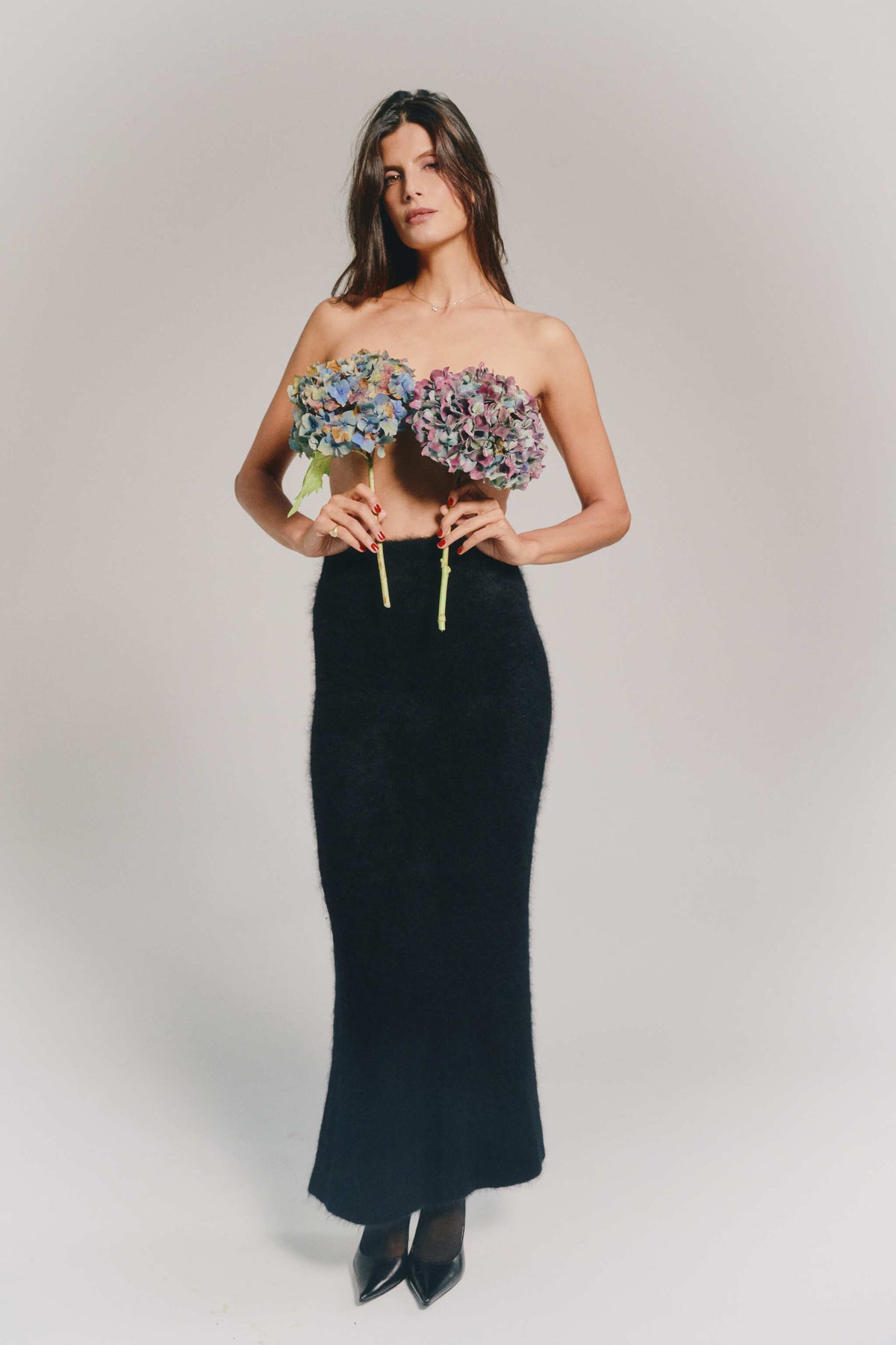 A woman modeling a long black cashmere skirt, paired with 2 matching colorful flowers. She is posing against a neutral light gray studio background.
