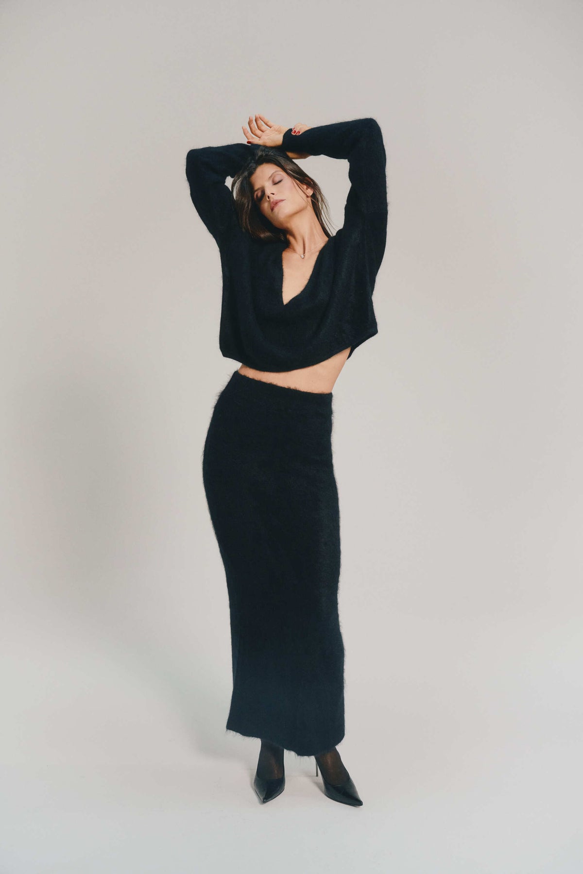 A woman modeling a black knit two-piece set consisting of a long-sleeve V-neck reversible cashmere knitwear and a matching high-waisted cashmere skirt, posed against a neutral light gray studio background.