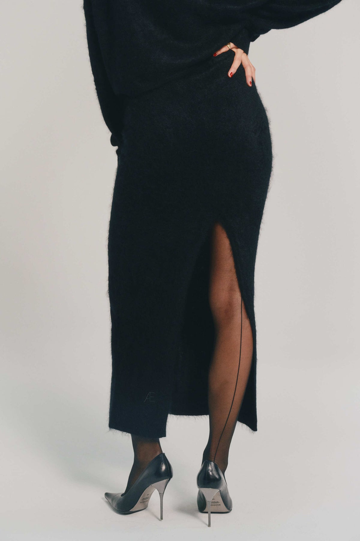 A model from the back wearing a black midi skirt in brushed cashmere with a high side slit, paired with black tights and black pointed-toe stiletto heels, against a light gray studio background.